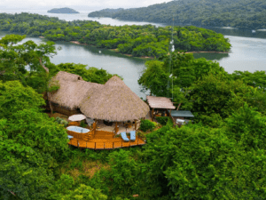 A thatched-roof hut with a wooden deck is surrounded by dense green foliage, overlooking a river and forested islands in the distance—an idyllic escape among top global travel destinations