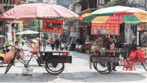 2 street vendors stand under a colorful umbrella beside a tricycle cart in an outdoor market, with signs in Chinese and tables in the background, inviting visitors to experience responsible tourism while supporting local livelihoods.