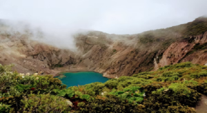 A tranquil blue lake sits in the center of a volcanic crater, with dramatic cliffs rising around its perimeter.