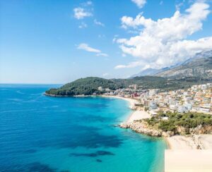 Aerial view of a coastal town with turquoise sea, sandy beaches, and buildings along the shoreline; this picturesque spot is one of the region’s hidden eco-friendly destinations, with hills and clouds visible in the background.